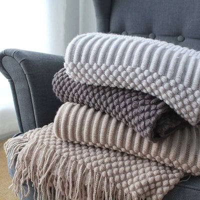 Stack of small knitted polyester nap blankets in neutral colors on a gray chair, featuring plush textures and fringe details.