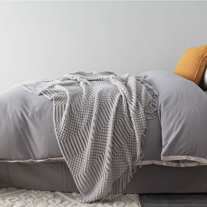 Small gray polyester fiber blanket draped over a bed with tassels, size 127x170cm, in a minimalist bedroom setting.