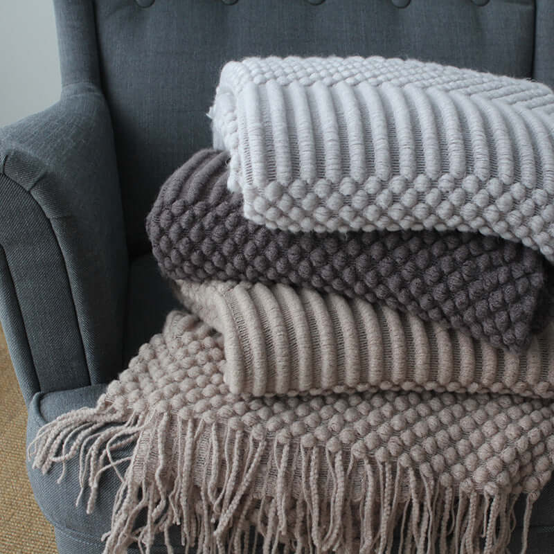 Stack of small polyester nap blankets in gray tones on a gray armchair, showcasing various textures and fringe details.