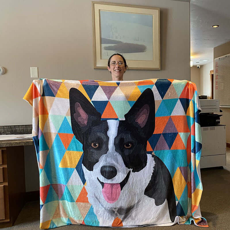 Woman holding a colorful flannel dog blanket featuring a vibrant cartoon-style corgi design in a cozy indoor setting.