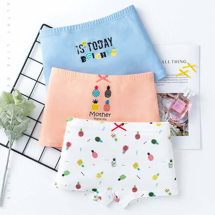 Set of three cotton girls' boxer underwear in pastel blue, peach, and white with fruit prints, displayed with books and decor.