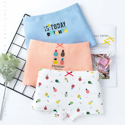 Set of three cotton girls' boxer underwear in pastel blue, peach, and white with fruit prints, displayed with books and decor.