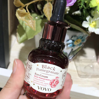 Hand holding a bottle of VOVO Black Pomegranate Eye-Cream with intricate label design, positioned against a backdrop of flowers.