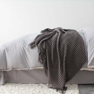 Small polyester nap blanket in dark gray draped artistically over a bed with light gray bedding and white rug.