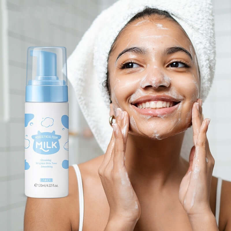 Woman with a towel on her head applying facial cleansing foam, smiling in bathroom with product visible